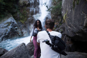 man in white carrying black sling bag taking photos at a girl carrying small black sacoche bag under a waterfall.