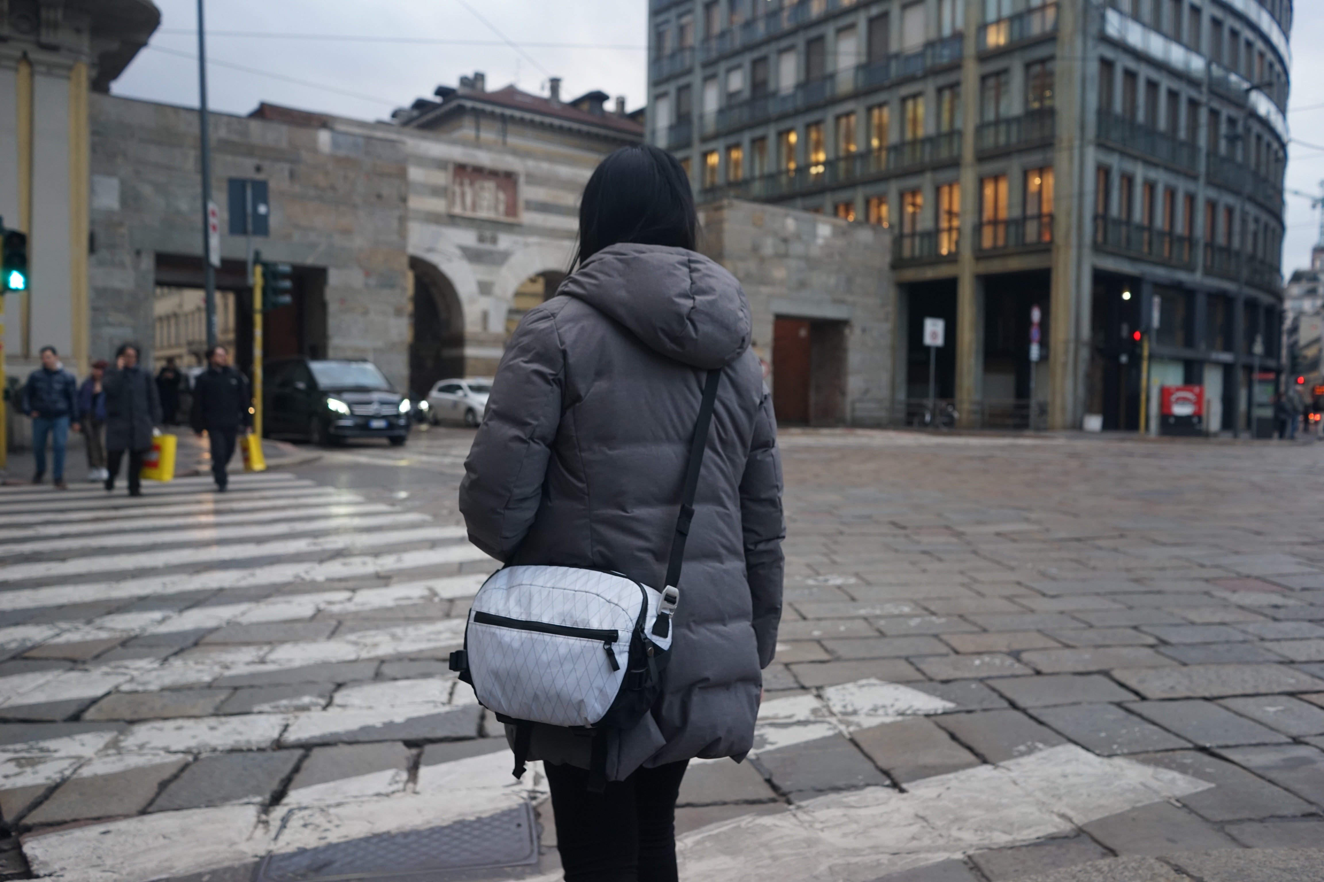 Girl in grey coat carrying white camera crossbody bag crossing a road in city